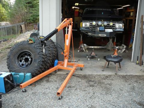 1991 Ford Bronco - A 1991 Ford Bronco is elevated in a garage, its front axle removed for maintenance, with a large tire and engine hoist nearby.