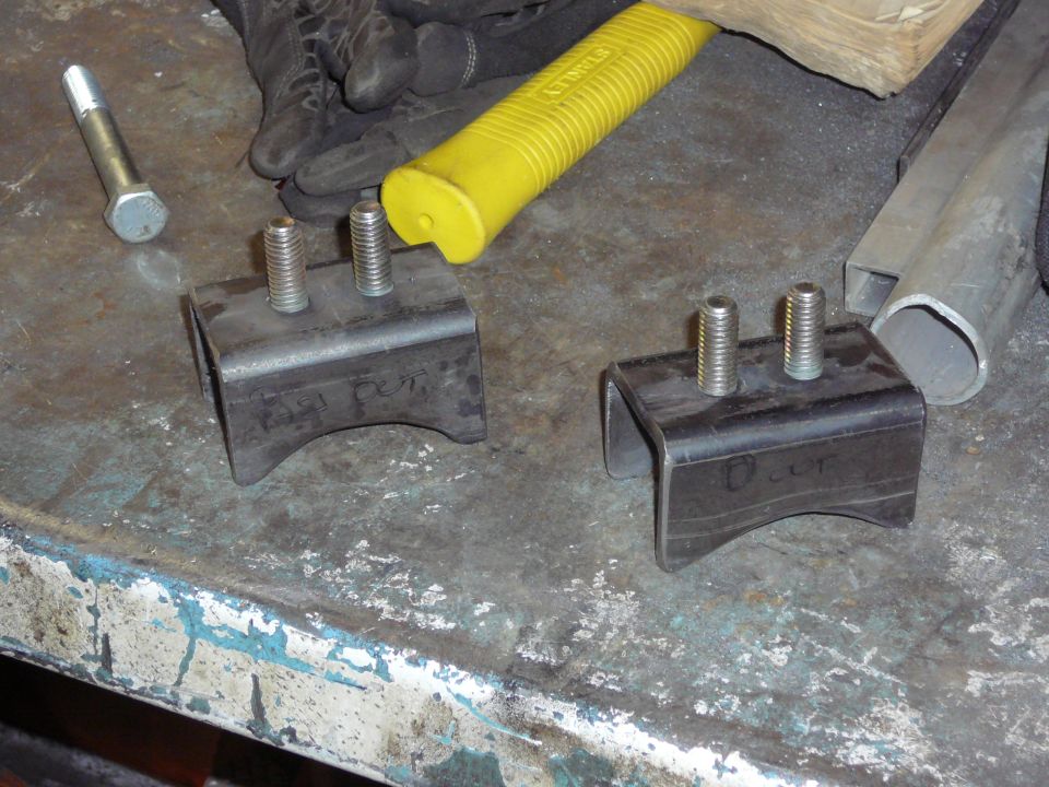 Two metal brackets with threaded bolts on a workbench next to a yellow hammer and gloves