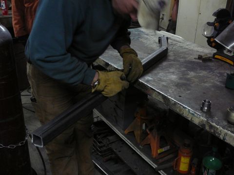1991 Ford Bronco - A person wearing a welding mask and protective gloves works on a metal frame at a workbench, with tools and equipment visible in the backgr…