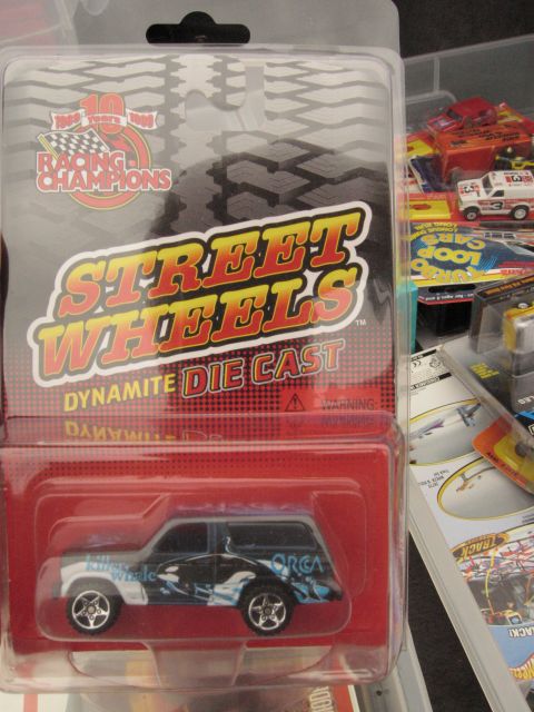 1986 Ford Bronco - A diecast toy SUV orca whale graphics is displayed in its packaging, alongside other toy vehicles.