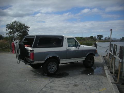 1991 Ford Bronco - This two-tone Ford Bronco is parked near a body of water, equipped with a rear-mounted spare tire and fuel cans.