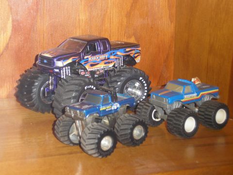 1995 Ford Bronco - A collection of toy monster trucks, including a purple Bigfoot truck and two blue Bigfoot trucks, are arranged on a shelf.