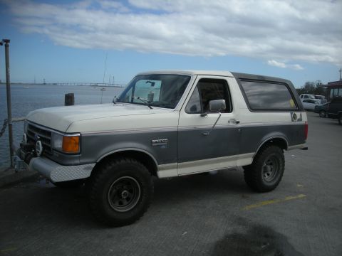 1991 Ford Bronco - A two-tone Ford Bronco is parked near a body of water under a cloudy sky.