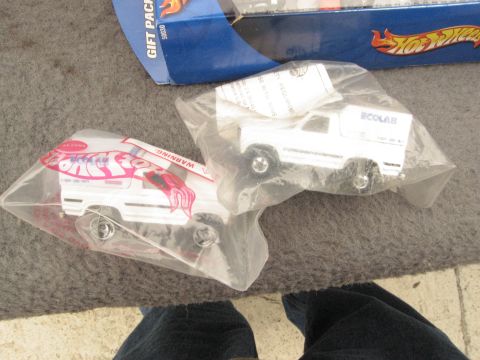 1986 Ford Bronco - Two white toy trucks are shown in their original packaging, with a Hot Wheels gift pack in the background.