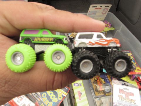 1986 Ford Bronco - A hand holds two toy monster trucks, one green and one white with flame decals, in front of a bin of other toys.