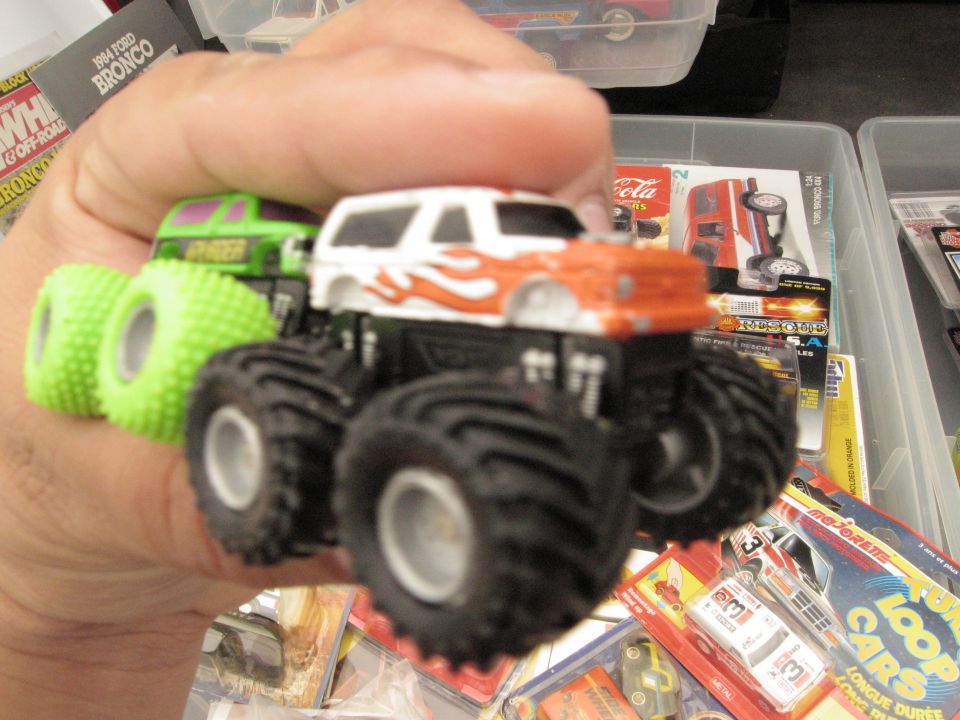 Hand holding a white and orange monster truck toy with green tires. A box labeled '1984 Ford Bronco' is visible in the background