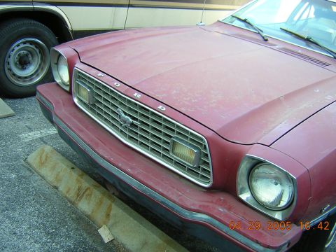 1975 Ford Mustang II - The front of a red 1975 Ford Mustang II is visible, with the grille and headlights in focus, next to another vehicle.