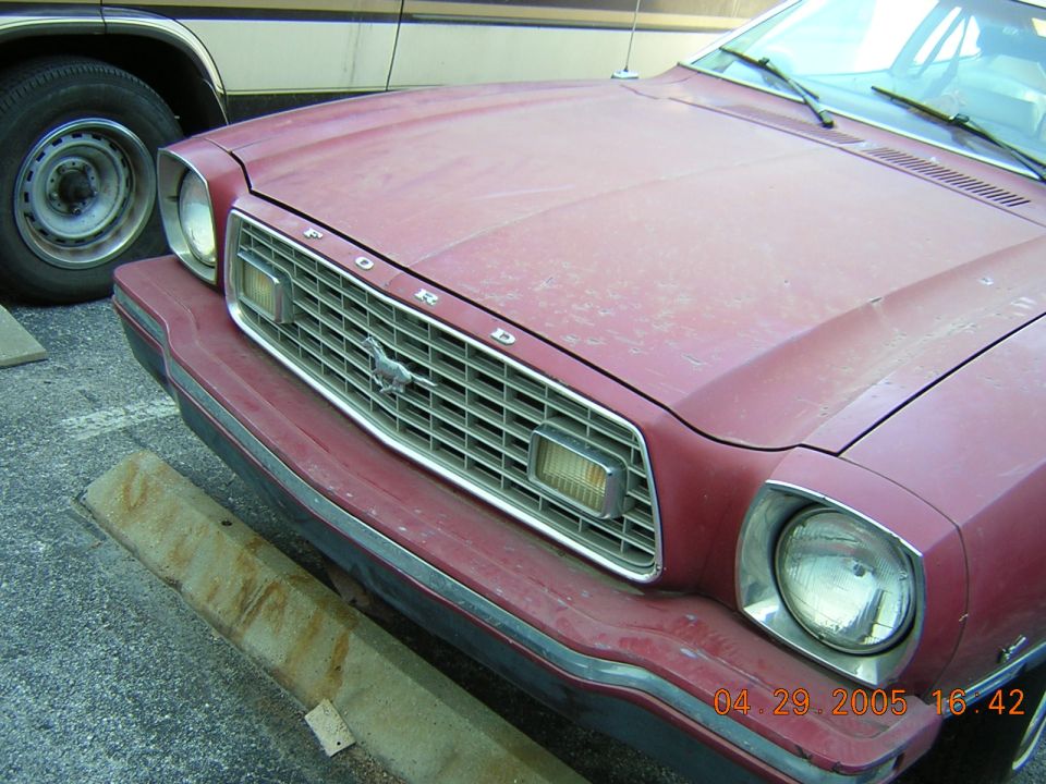 Red 1975 Ford Mustang II front grille with a horse emblem and 'FORD' lettering, showing a faded paint job and a portion of another vehicle on the left