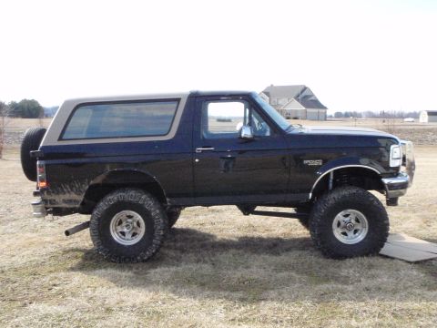1996 Ford Bronco - The side profile of a black 1996 Ford Bronco is shown parked on a dry grassy field. The vehicle has a tan top and large off-road tires.