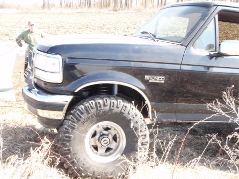 1996 Ford Bronco - A black Ford Bronco is parked in a dry, grassy field with a person standing behind it. The front passenger side of the vehicle, including t…