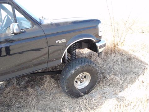 1996 Ford Bronco - A black Ford Bronco is shown from the side, with a lifted suspension and large off-road tires. The vehicle is parked on dry grass with some…