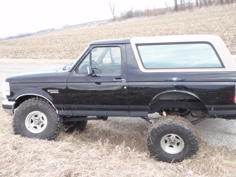 1996 Ford Bronco - The side view of a black 1996 Ford Bronco with a tan roof and large off-road tires is parked on a gravel road next to a dry field.
