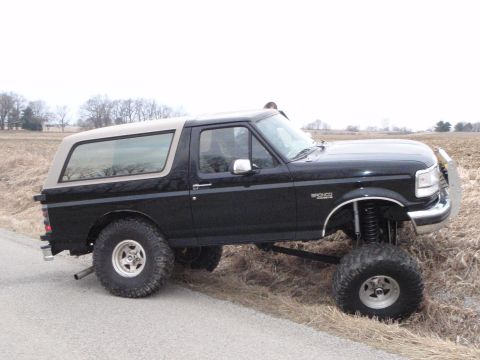 1996 Ford Bronco - A lifted black 1996 Ford Bronco with a tan removable top is parked on the side of a dirt road next to a dry field.