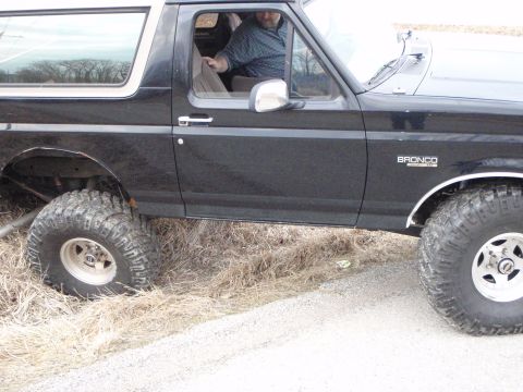 1996 Ford Bronco - The side view of a black Ford Bronco with a tan top is parked on a dirt road with dry grass. The vehicle has large off-road tires and the B…
