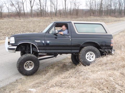 1996 Ford Bronco - A lifted black 1996 Ford Bronco with a tan roof is parked on the side of a dirt road, with dry grass and trees in the background. A man is…