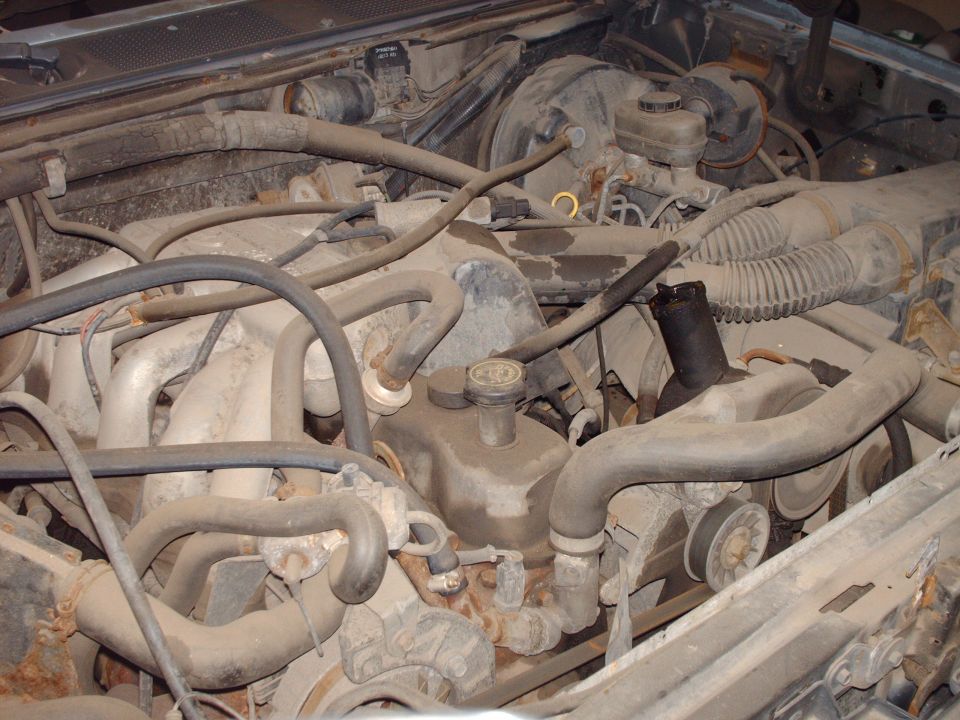 Dusty engine bay with hoses, intake manifold, and engine block visible.