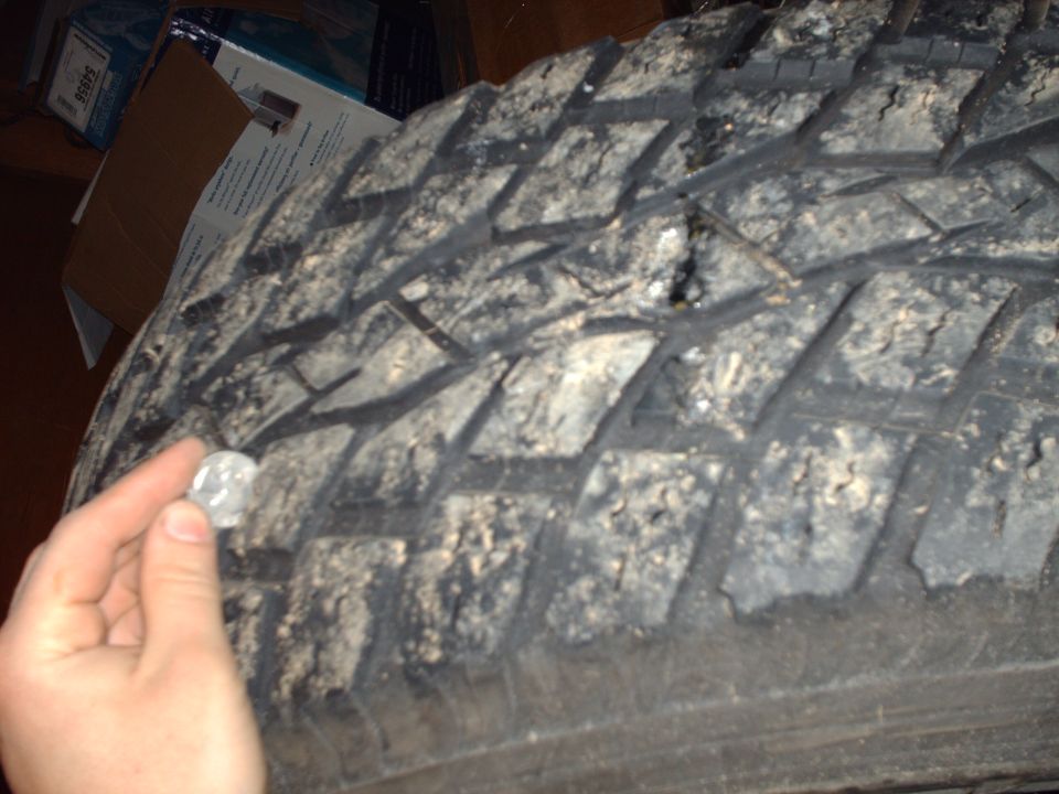 Close-up of a dirty tire tread with a hand holding a coin near a puncture.
