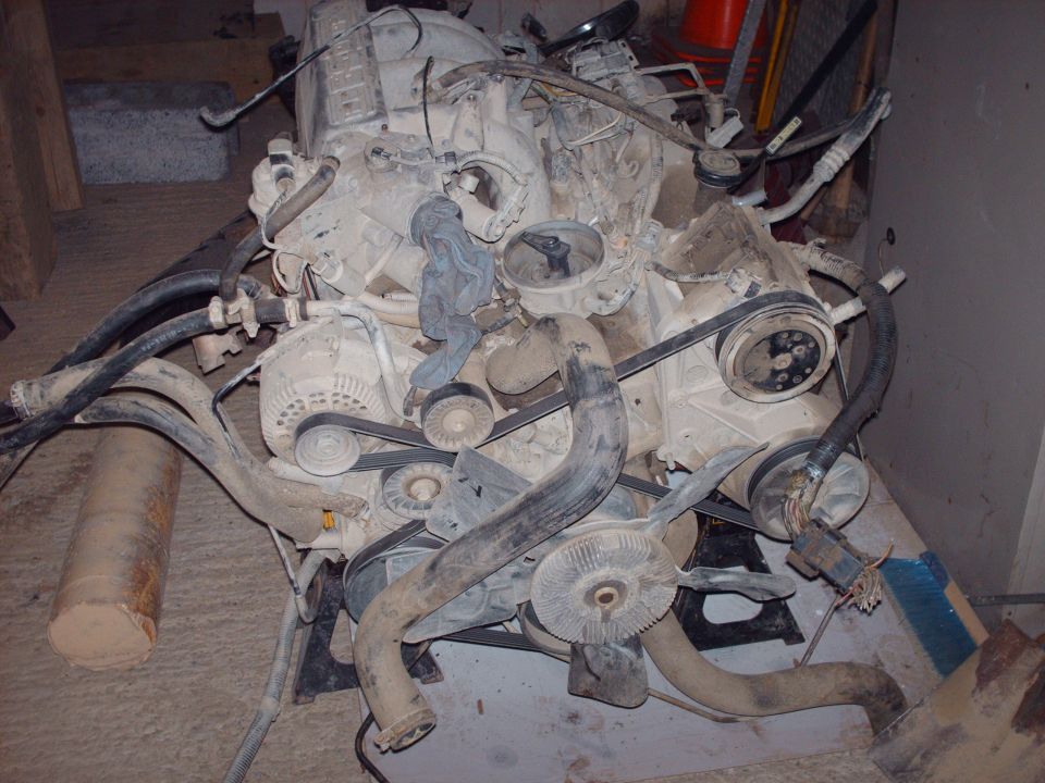 Disassembled V-type engine with hoses, belts, and pulleys on a concrete floor.