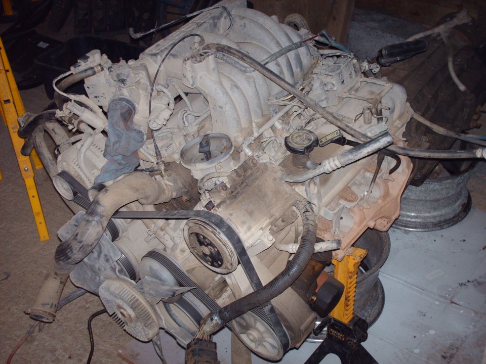 Dusty V8 engine with intake manifold, belts, hoses, and wires, supported by a yellow jack.
