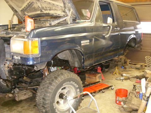 1988 Ford Bronco - A dark blue Ford Bronco is in a garage with its hood open and the front end disassembled. The vehicle is raised on jacks with various tools…