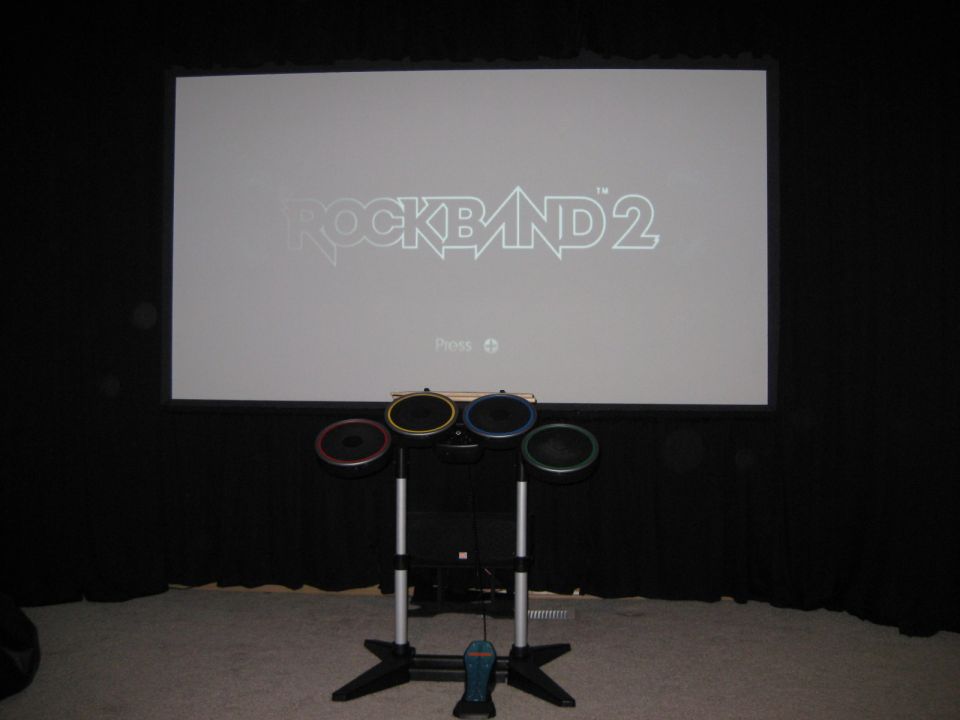 Rock Band 2 logo displayed on a projector screen with a drum set and pedal in front
