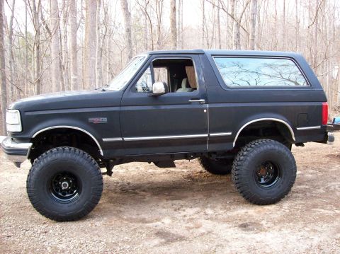 1992 Ford Bronco - This lifted 1992 Ford Bronco features oversized tires and a dark exterior, presented in a side profile against a wooded background.