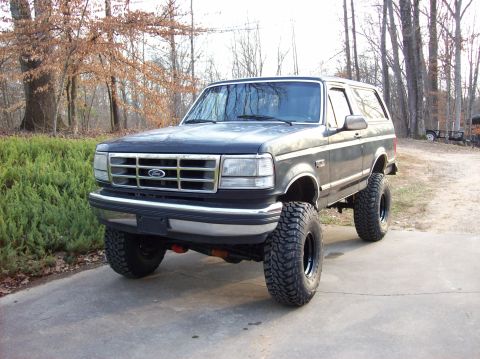 1992 Ford Bronco - A lifted 1992 Ford Bronco sits on a paved surface with trees in the background.