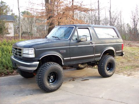 1992 Ford Bronco - This lifted 1992 Ford Bronco features large off-road tires and a dark gray exterior, parked on a paved surface with a wooded background.