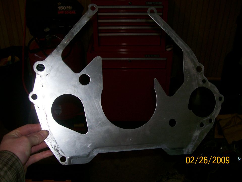 Metal transmission bell housing adapter plate held by a hand, with a red toolbox in the background