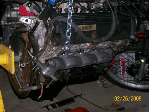 1995 Mercury Grand Marquis - An engine assembly, likely removed from a vehicle, is suspended by a chain, with an exhaust manifold connected.