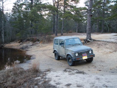 1987 Suzuki Samurai - A gray Suzuki Samurai is parked on a dirt path surrounded by trees and a body of water.