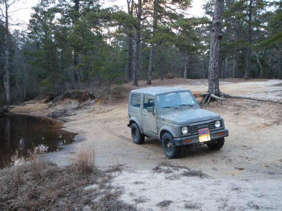 Gray Suzuki Samurai parked on dirt near trees and water