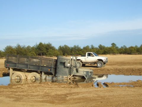 1987 Suzuki Samurai - A military flatbed truck is stuck in a muddy area, with a white pickup truck visible behind it.