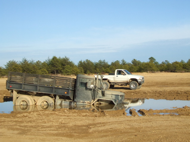 Military flatbed truck stuck in mud, white pickup truck in background