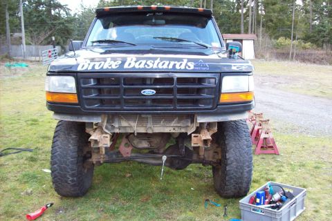 1995 Ford Bronco - This black Ford truck, missing its front bumper and bearing custom text, is elevated on jack stands.