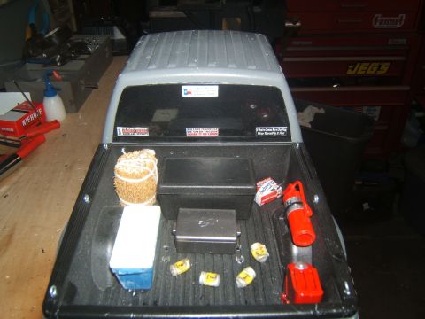 1988 Ford Bronco - The bed of a gray pickup truck is filled with various items including a cooler, a bundle of hay, a fire extinguisher, and several beer cans…