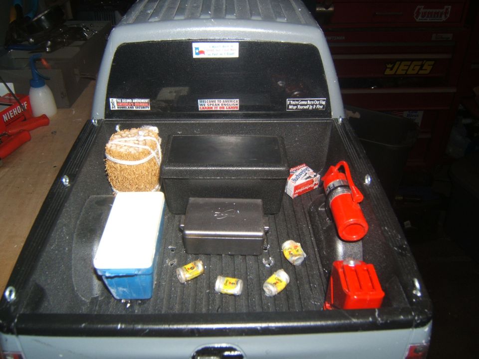 Pickup truck bed with cooler, hay, toolboxes, fire extinguisher, and cans. Rear window with stickers.
