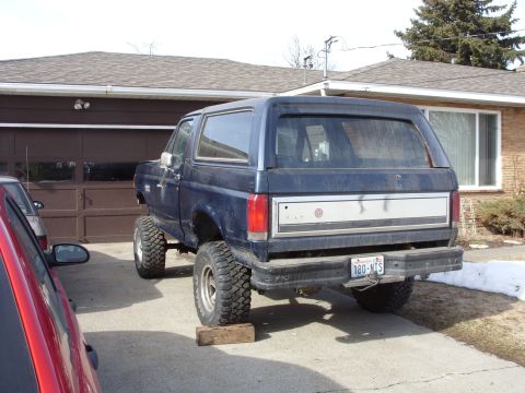 1988 Ford Bronco - The rear driver-side quarter view of a lifted, dark blue Ford Bronco is shown parked in a driveway in front of a garage. The vehicle is rai…