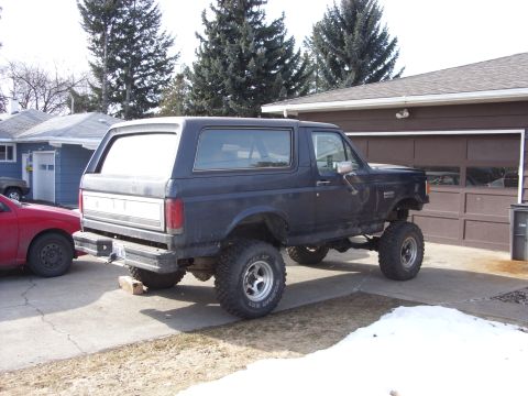 1988 Ford Bronco - A lifted, dark blue Ford Bronco is parked in a driveway next to a red car. The Bronco has large off-road tires and is raised significantly…