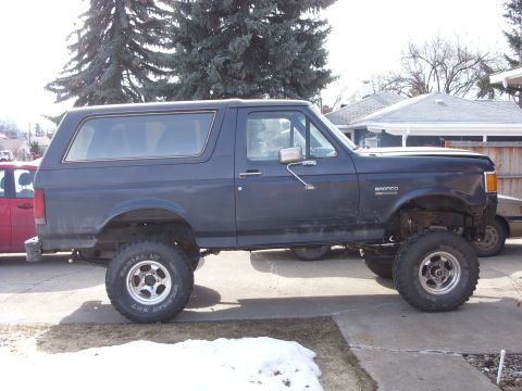 1988 Ford Bronco - A dark blue Ford Bronco is parked on a concrete driveway with snow on the ground. The vehicle is lifted with large off-road tires and appea…