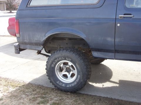 1988 Ford Bronco - The rear driver's side quarter panel and wheel of a dark blue Ford Bronco are shown. The vehicle has a lifted suspension and large off-road…