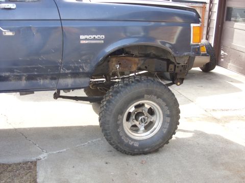 1988 Ford Bronco - The driver's side of a dark blue Ford Bronco is shown lifted with large off-road tires. The vehicle has a "BRONCO XLT" badge visible on the…