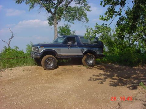 1986 Ford Bronco - 1986 Ford Bronco climbs a dirt embankment under a sunny sky with trees overhead.