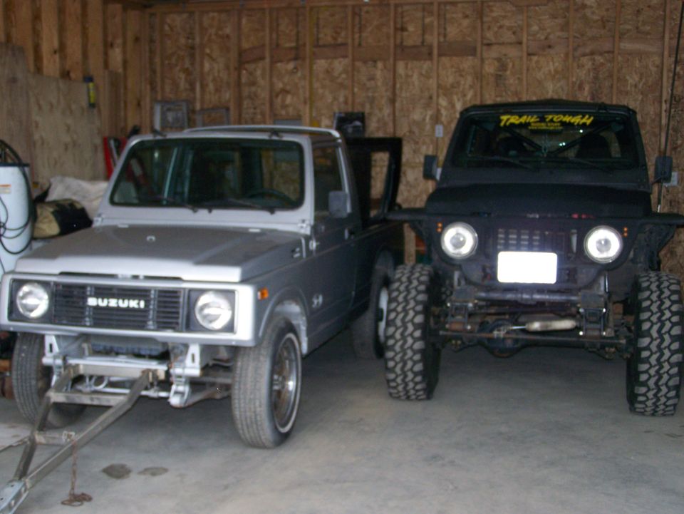 Two Suzuki Samurai vehicles in a garage, one silver convertible and one black off-road model.