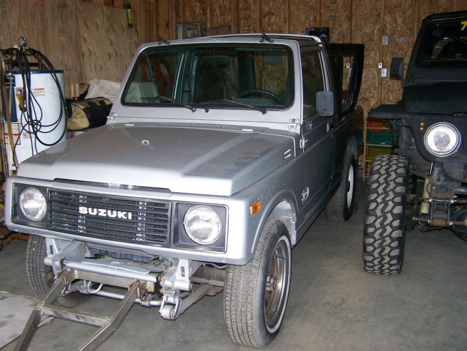 Silver 1988 Suzuki Samurai in a garage with a custom front bumper, parked next to a black Jeep.