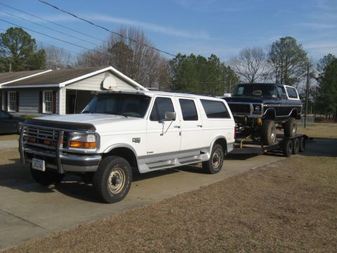 1978 Ford Bronco - A white SUV tows a lifted dark blue 1978 Ford Bronco on a trailer down a driveway.