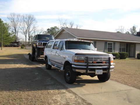 1978 Ford Bronco - A white SUV tows a blue pickup truck on a trailer in front of a house.