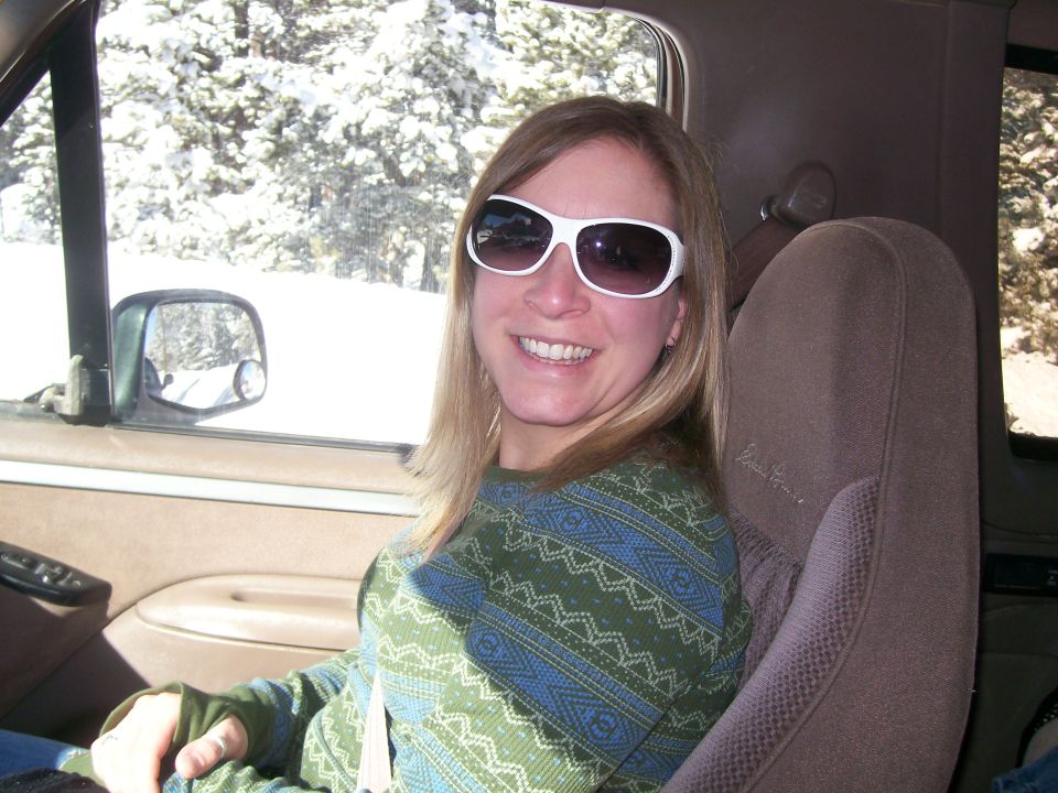 Woman in sunglasses smiles in the driver's seat of a Ford Bronco, snow-covered trees outside.