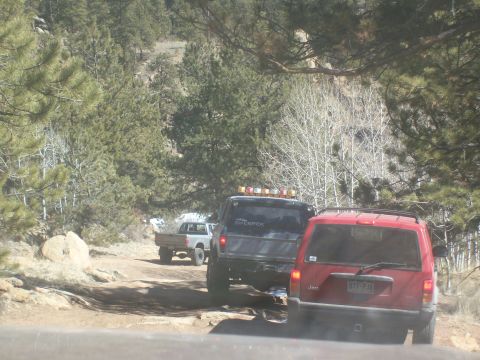 1993 Ford Bronco - A red Jeep Cherokee is parked on a dirt road behind a gray pickup truck, surrounded by pine trees. The Jeep has a license plate that reads…