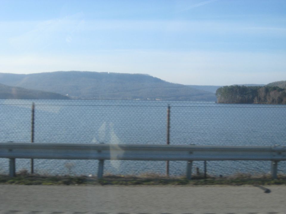 Lake behind a chain-link and guardrail fence with forested hills